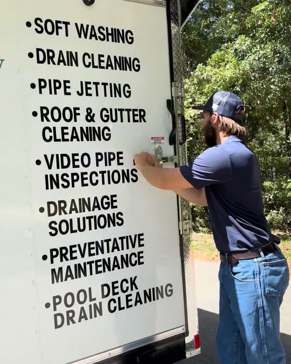 A technician opening the trailer, its door listing the eight services the rig carries, from soft washing to pool deck drain cleaning