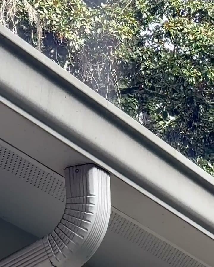 A downspout and its elbow at the corner of a house under the gutter, with rinse water still misting off the roof edge