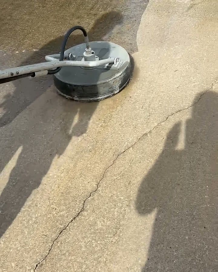Surface cleaner mid-pass on a concrete driveway, the cleaned strip bright against the untouched grey beside it