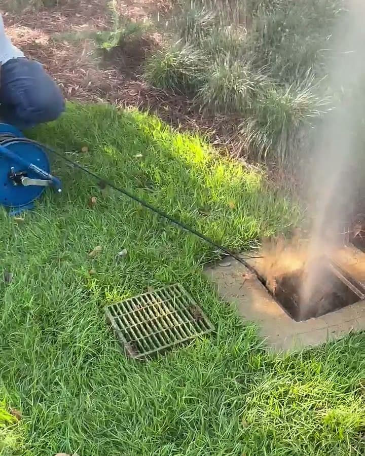 Jetter line fed from a hose reel into an open yard drain inlet, water and loosened debris blasting back out as the line clears